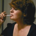 Still from the film "Souris, tu m'inquiètes" featuring a white woman with short brown hair holding a cigarette in her mouth.