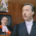 Still from the film "Conspiracy of Silence" featuring a judge and a lawyer, both middle aged white men with moustaches, in a courtroom.