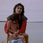 Still from the film "A Safe Distance" featuring an Indigenous woman sitting with her young child in her lap with a lake in the background.