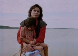 Still from the film "A Safe Distance" featuring an Indigenous woman sitting with her young child in her lap with a lake in the background.