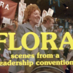 A still from the film "Flora" featuring the title of the film in yellow text. In the background Flora MacDonald, a middle-aged white woman with curly light brown hair wearing a grey suit, is standing and smiling at a packed campaign event.