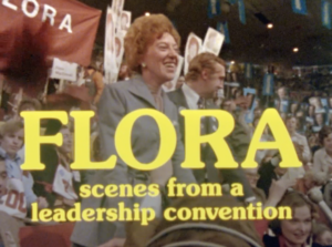 A still from the film "Flora" featuring the title of the film in yellow text. In the background Flora MacDonald, a middle-aged white woman with curly light brown hair wearing a grey suit, is standing and smiling at a packed campaign event.