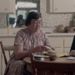 Still featuring Margaret Laurence, an older white woman with short grey hair and glasses wearing a floral dress, sitting at a kitchen table and opening letters.