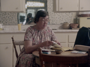 Still featuring Margaret Laurence, an older white woman with short grey hair and glasses wearing a floral dress, sitting at a kitchen table and opening letters.