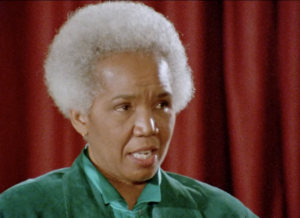 Still from the film "No Way! Not Me" featuring Rosemary Brown, an older Black woman with white cropped hair wearing a teal shirt, speaking on a stage.