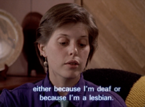 Still from the film "Toward Intimacy" featuring Amethya Weaver, one of the film's subjects, talking about her deaf and lesbian identity. She is a white woman with short brown hair wearing a purple shirt and earrings.