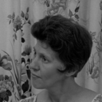 Still from the film "A Woman's Place" featuring a white woman listening to another woman talking. She has short brown hair. Behind her is a floral curtain.