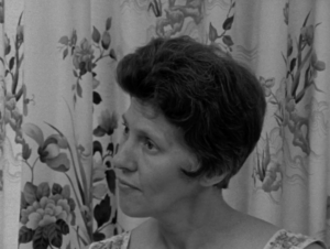 Still from the film "A Woman's Place" featuring a white woman listening to another woman talking. She has short brown hair. Behind her is a floral curtain.