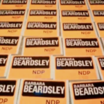 Still from the film "Campaign" featuring yellow and black placards with "Barbara Beardsley NDP" written on them.