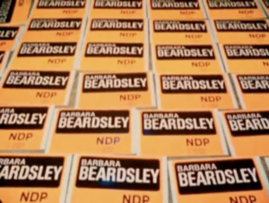 Still from the film "Campaign" featuring yellow and black placards with "Barbara Beardsley NDP" written on them.