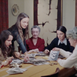 Still from the film "They Called Us 'Les Filles du Roy'" featuring an intergenerational group of Québecois women sitting at a table and looking at old photographs.