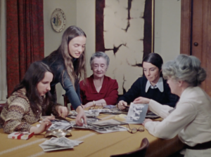 Still from the film "They Called Us 'Les Filles du Roy'" featuring an intergenerational group of Québecois women sitting at a table and looking at old photographs.