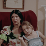 Still from the film "Our Dear Sisters" featuring Alanis Obomsawin, an Indigenous women wearing braids and a dress, sitting in an arm chair and talking to the camera, with her young daughter in her lap.