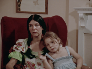 Still from the film "Our Dear Sisters" featuring Alanis Obomsawin, an Indigenous women wearing braids and a dress, sitting in an arm chair and talking to the camera, with her young daughter in her lap.