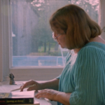 Still from the film "Firewords" featuring Louky Bersianik, a middle-aged white woman with light brown hair wearing a teal blouse, writing at her desk.