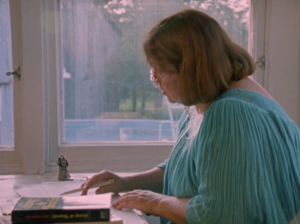 Still from the film "Firewords" featuring Louky Bersianik, a middle-aged white woman with light brown hair wearing a teal blouse, writing at her desk.