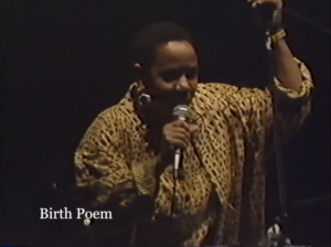 Screenshot of Lillian Allen, a Black woman with short hair wearing a yellow and brown blouse, performing Birth Poem on stage.