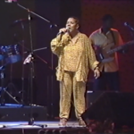 Still of Lillian Allen, a Black woman with short hair wearing a yellow jumpsuit, singing "I Fight Back" on stage.