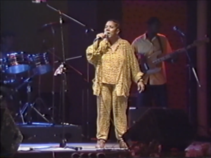 Still of Lillian Allen, a Black woman with short hair wearing a yellow jumpsuit, singing "I Fight Back" on stage.