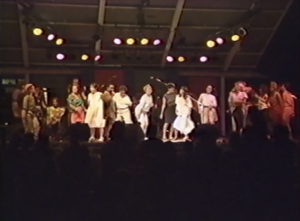 Still of Lillian Allen performing The Subversives. She is joined on stage by many women, who are all dancing.