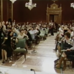 Still from footage of the constitutional conference in 1981 featuring a large group of white feminists sitting in a room in Parliament. Many of them are raising their hands to vote on a motion.