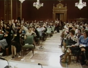 Still from footage of the constitutional conference in 1981 featuring a large group of white feminists sitting in a room in Parliament. Many of them are raising their hands to vote on a motion.