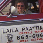 Still from the film "Laila" featuring Laila, a white woman with curly hair wearing sunglasses, in a red car with a sign that says "Laila Paattinen Drywall Taping" on the side of the car.