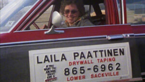 Still from the film "Laila" featuring Laila, a white woman with curly hair wearing sunglasses, in a red car with a sign that says "Laila Paattinen Drywall Taping" on the side of the car.