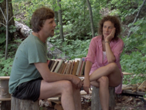 Still of filmmaker Michael Rubbo and Margaret Atwood sitting on tree stumps and talking at a campsite beside a row of her books.