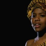 Still from the film "Speak It" featuring a young black woman wearing a yellow patterned head wrap looking into the distance against a black background.