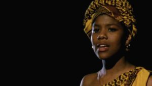 Still from the film "Speak It" featuring a young black woman wearing a yellow patterned head wrap looking into the distance against a black background.