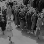 Still from the film "Democracy à la Maude" featuring a black and white archival photograph of white women and children marching in the street.