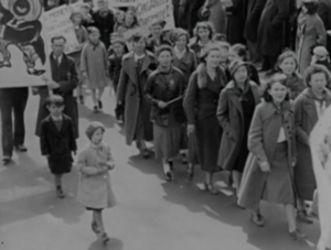 Still from the film "Democracy à la Maude" featuring a black and white archival photograph of white women and children marching in the street.