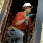 Still from the film "Building Futures" featuring a white woman wearing a red shirt, blue jeans, and a white hard hat drilling a door frame.