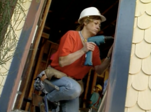 Still from the film "Building Futures" featuring a white woman wearing a red shirt, blue jeans, and a white hard hat drilling a door frame.