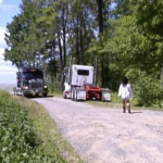 Still from the film "5'2'80 000 LBS" featuring a woman walking behind two trucks on a dirt road lined with trees.