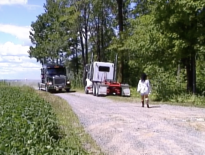 Still from the film "5'2'80 000 LBS" featuring a woman walking behind two trucks on a dirt road lined with trees.