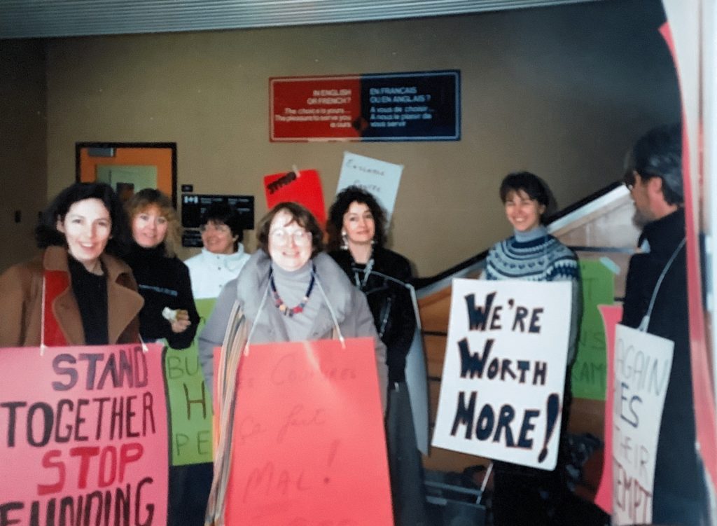 Group of women with signs demonstrating in a hall. "We're Worth More!"