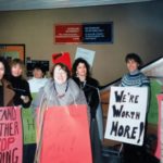 Group of women with signs demonstrating in a hall. "We're Worth More!"