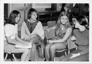 Group of four women sitting in chairs, talking at a women's conference held in Sault Ste. Marie i the 1970s. One is holding a notebook.