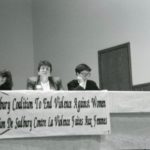 Three women sitting behind a table with a banner that says "The Sudbury Coalition to End Violence Against Women / La Coalition De Sudbury Contre La Violence Faites Aux Femmes"