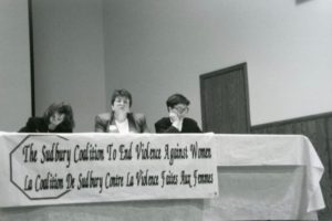 Three women sitting behind a table with a banner that says "The Sudbury Coalition to End Violence Against Women / La Coalition De Sudbury Contre La Violence Faites Aux Femmes"