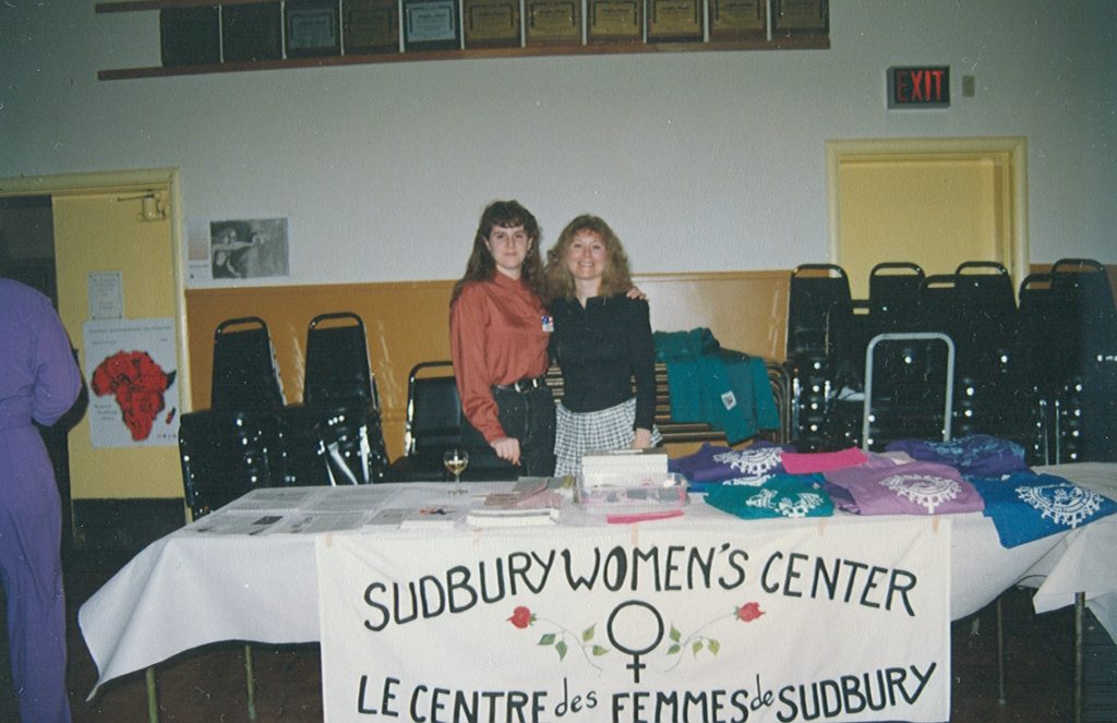 Two women behind a table with shirts and papers on it. Banner in front of table that says "Sudbury Women's Center / Le Centre des Femmes de Sudbury"