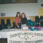 Two women behind a table with shirts and papers on it. Banner in front of table that says "Sudbury Women's Center / Le Centre des Femmes de Sudbury"