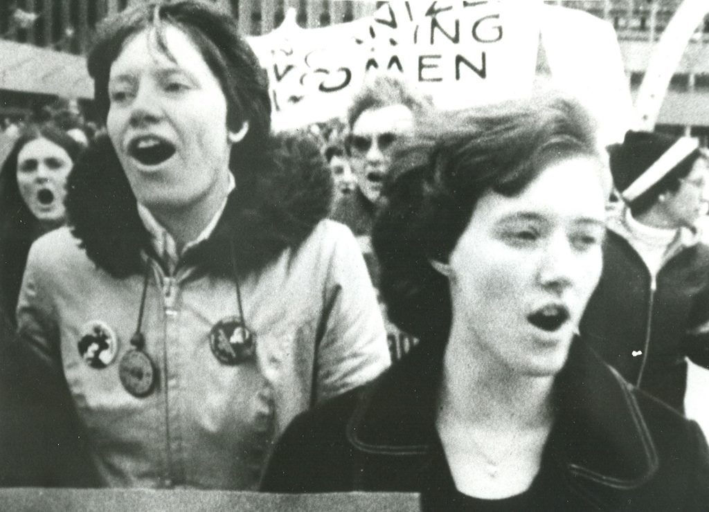 Group of women marching, banner in the background.