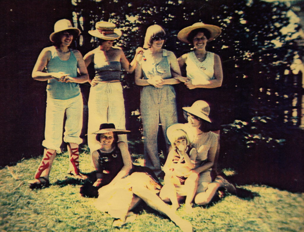 Scanned photograph of six members of Isis collective in a garden. Four members are standing and two are sitting on the ground.