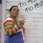 Still from the short film "Unnatural Causes" depicting Lillian Allen, a black woman with short black hair wearing colourful overalls, speaking to the camera and writing the words "no rest, no rest" on a white brick wall.