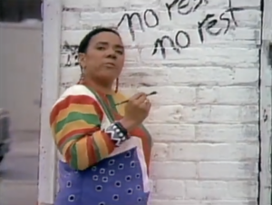 Still from the short film "Unnatural Causes" depicting Lillian Allen, a black woman with short black hair wearing colourful overalls, speaking to the camera and writing the words "no rest, no rest" on a white brick wall.