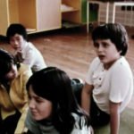 Still from the film "Starting from Nina" depicting archival footage of four kids sitting on the floor of a classroom and talking.