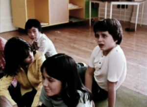 Still from the film "Starting from Nina" depicting archival footage of four kids sitting on the floor of a classroom and talking.